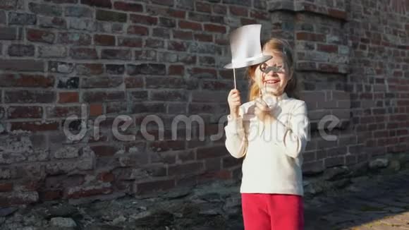 小女孩拿着纸胡子和帽子在砖墙旁边的棍子上视频的预览图