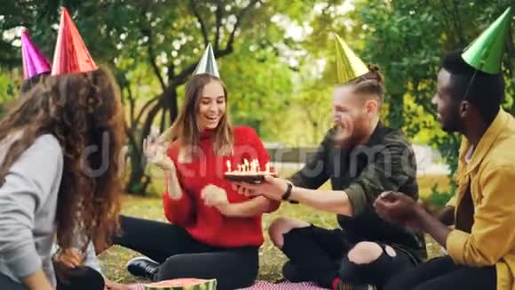 戴着派对帽的漂亮女孩在公园野餐时和朋友们一起庆祝生日许愿在蛋糕上吹蜡烛视频的预览图