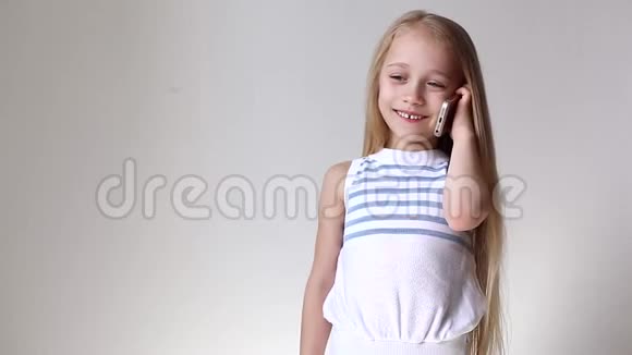 一个微笑的金发小女孩穿着白色背景的休闲T恤在家里打电话视频的预览图