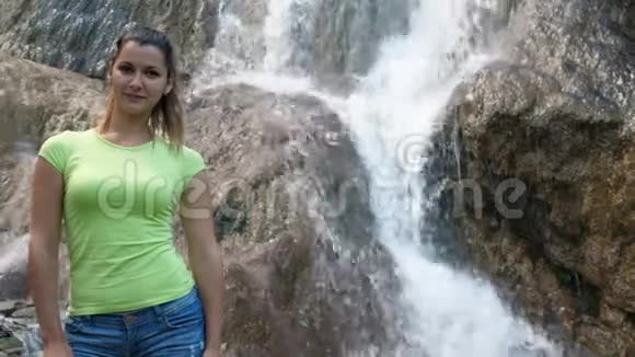 一个女人站在瀑布的背景上快速的瀑布沿着岩石流下来一个美丽的微笑女孩旁边视频的预览图
