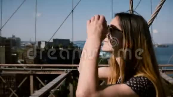 镜头跟随美丽快乐的欧洲女孩戴着老式太阳镜金色的头发走在布鲁克林大桥4K视频的预览图