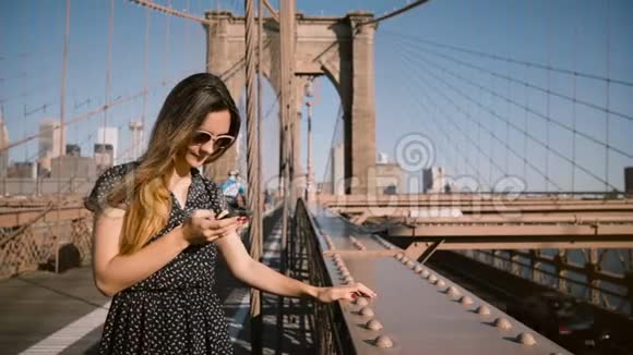 美丽微笑的欧洲博主女士戴着时尚太阳镜在布鲁克林大桥使用智能手机应用程序走开了4K视频的预览图