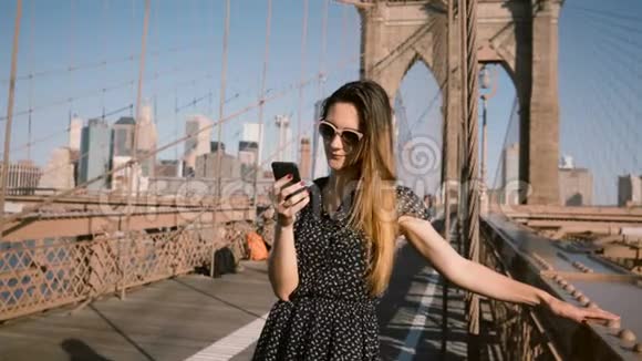 快乐的年轻白种人女士戴着老式太阳镜在电话里交谈在布鲁克林大桥4K的风中吹着头发视频的预览图