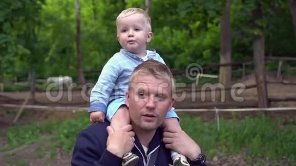 儿子坐在父亲脖子上在春天阳光明媚的公园里散步父亲天特写镜头视频的预览图