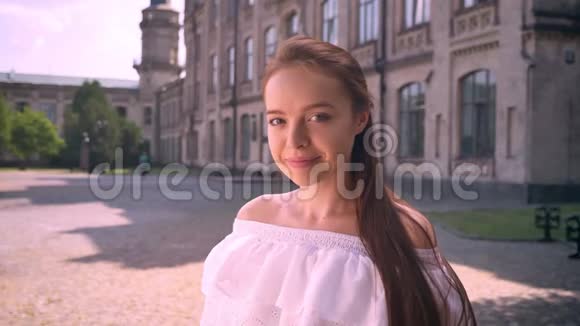 迷人的年轻女子看着镜头微笑站在街上背景是建筑阳光明媚的室外视频的预览图