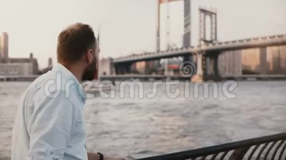 成功的欧洲商人站在纽约曼哈顿大桥附近令人惊叹的日落河上红色游艇经过4K视频的预览图