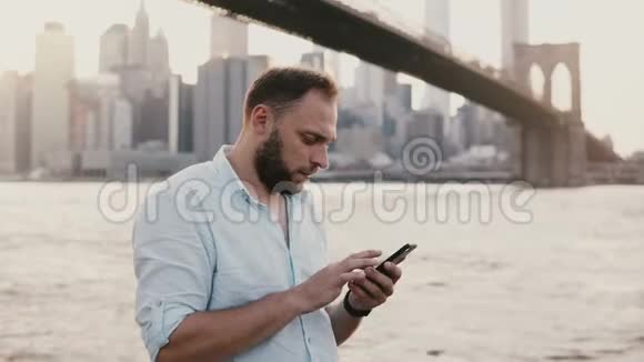 快乐的白种人自由职业企业家在智能手机上打字信息微笑着靠近布鲁克林大桥纽约市4K视频的预览图