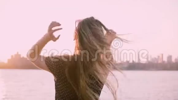 后观欢乐女独自站着看着江岸上令人惊叹的日落摸着头发和手臂张开的慢动作视频的预览图