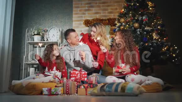 幸福的家庭有四个人他们带着火花微笑着看着圣诞树脚下的摄像机视频的预览图