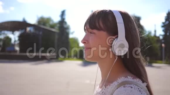 一个女孩带着耳机沿着城市街道走听音乐和微笑穿着白色长裙长发的女孩视频的预览图