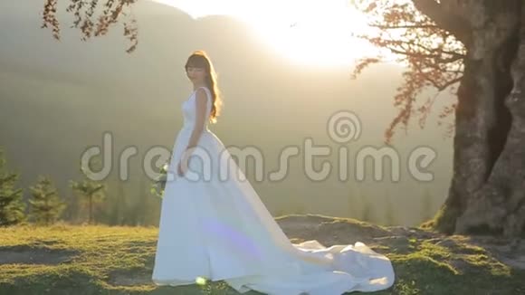 新娘在日落时穿着婚纱在树和山的背景下太阳从外面下山视频的预览图