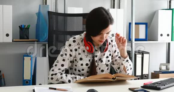 在现代办公室戴着红色耳机的女人用铅笔在笔记本上写字视频的预览图