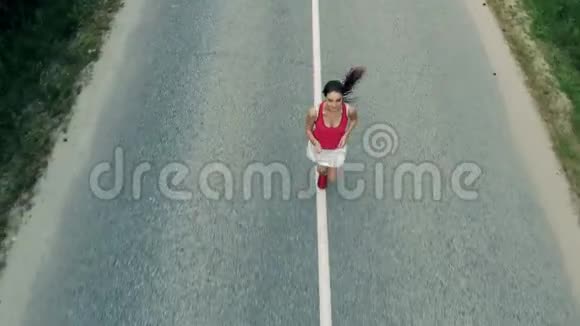 在森林中沿着高速公路奔跑的一位漂亮的年轻女子的正面俯视空中飞行视频的预览图