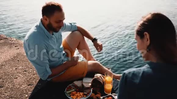 年轻的幸福夫妇坐在海边吃饭早上在水边野餐的男人和女人视频的预览图