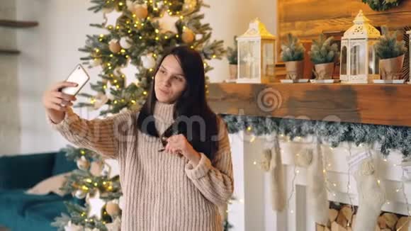 美丽的年轻女子在圣诞节时正站在壁炉壁炉架和新壁炉架附近用智能手机自拍视频的预览图