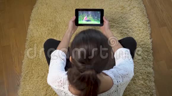 一个女人用平板电脑观看屏幕上的运动训练而坐在家里的地毯上视频的预览图