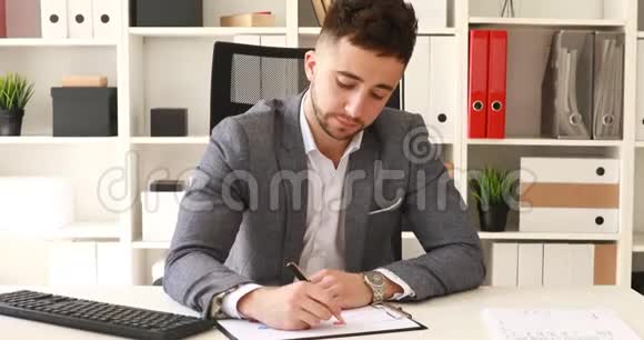 穿着灰色夹克的商人坐在白色办公室的桌子上写着文件视频的预览图