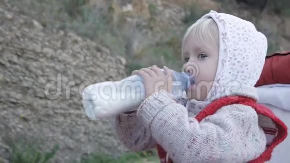 穿着帽衫和红色针织背心的小女孩在婴儿车里坐着塑料瓶喝牛奶或奶制品视频的预览图