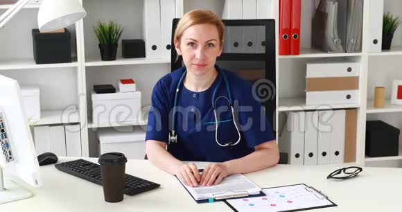 穿着蓝色衣服的医生坐在工作场所看着摄像机视频的预览图