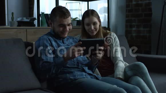 年轻的一对带平板电脑的微笑情侣在沙发上放松视频的预览图