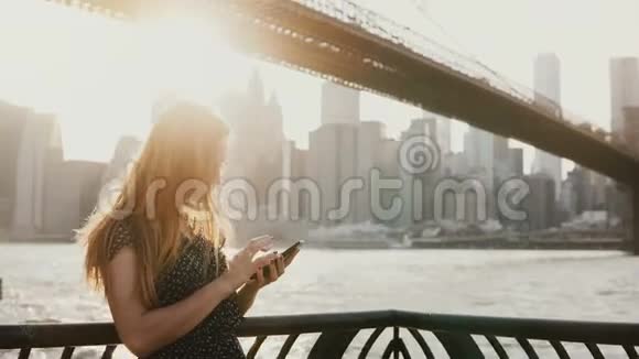 美丽的女孩戴着长长的太阳镜在纽约布鲁克林大桥附近的日落河码头使用智能手机应用程序视频的预览图