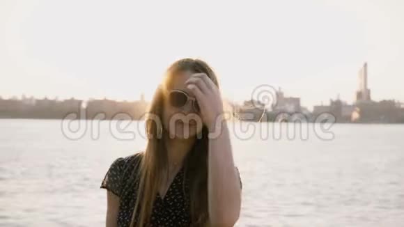 快乐休闲的白种人女孩戴着时尚的太阳镜在相机上摆姿势纽约日落天际线和背景4K的河流视频的预览图