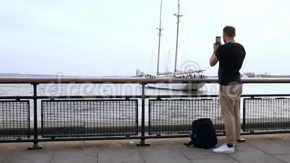 年轻英俊的男人背包站在码头上在智能手机上拍摄摩托艇的照片视频的预览图