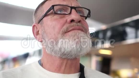 机场一位留着灰色胡须和眼镜的成熟男人的亲密肖像养老金领取者抬头看着航班视频的预览图