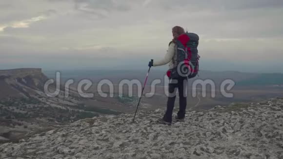 年轻的徒步旅行者女人带着背包和登山杆站在岩石的边缘视频的预览图