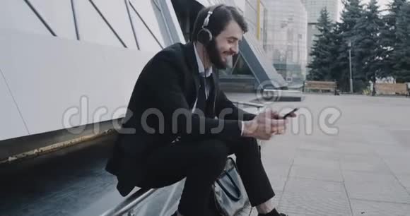 年轻英俊的商人穿着西装用手机在商务中心前面听音乐视频的预览图