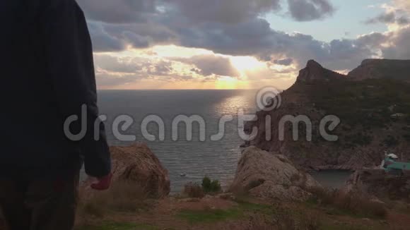 身穿夹克衫头戴帽子的人在五彩缤纷的夕阳下走到靠近海岸的岩石悬崖边视频的预览图