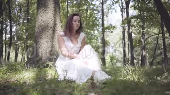 画中一个可爱的年轻女孩留着长长的黑发身穿白色夏季时装长裙坐在一棵树下视频的预览图