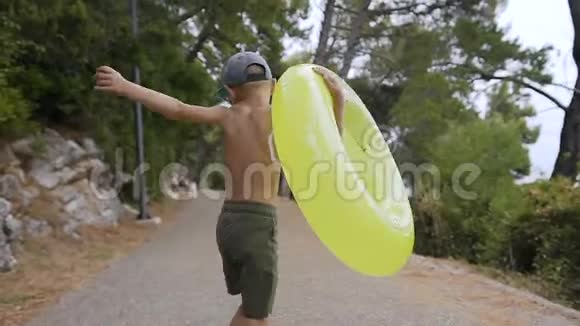 回头看一个穿着短裤的小男孩带着充气子欢快地沿着公园跑到海滩在阳光明媚的夏天视频的预览图