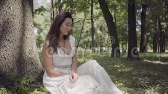 可爱的年轻女孩的肖像长着一头深色长发穿着一件白色的夏季时装长裙坐在树下视频的预览图