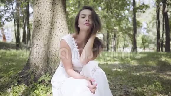 可爱的年轻女孩的肖像长着一头深色长发穿着一件白色的夏季时装长裙坐在树下视频的预览图