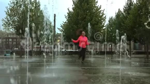 年轻的混血女人在城市公园喷泉附近的雨中奔跑慢镜头视频的预览图
