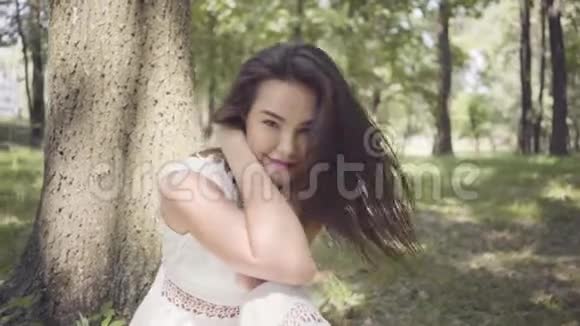 一个可爱的小女孩长着深色长发穿着一件白色的夏装坐在树下视频的预览图