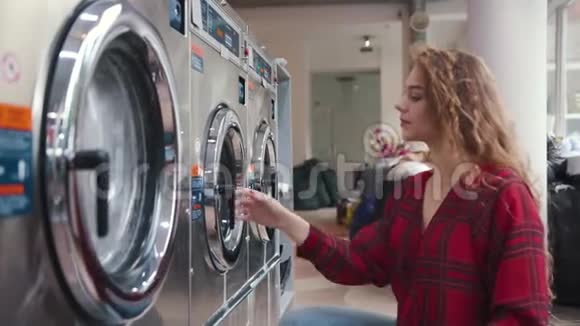 漂亮的年轻女人有着红色的卷发把衣服放进洗衣店里的洗衣机里穿着格子衬衫视频的预览图