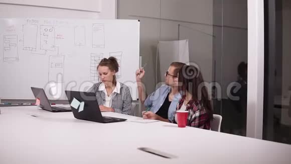 戴眼镜的年轻人和两个随意坐在办公室桌子上的女人在创意工作场所上面有笔记本电脑男子视频的预览图