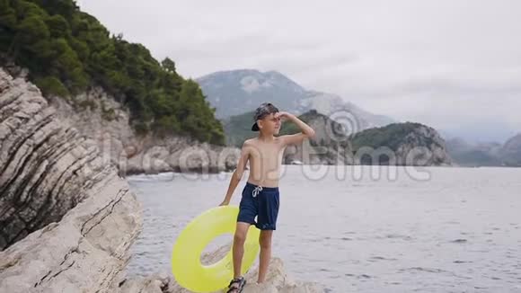 漂亮的小男孩穿着短裤和带充气黄色圆圈的帽子站在悬崖上举起手臂视频的预览图
