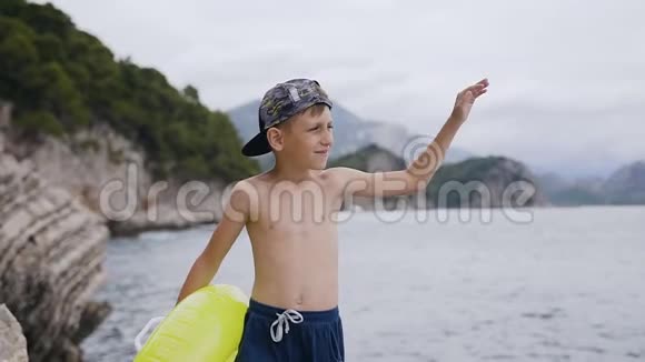 可爱的小男孩站在岩石上在日落时眺望大海一个带充气戒指的可爱男孩的肖像视频的预览图