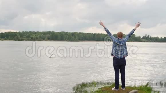 一个年轻男子站着举起双手站在美丽的河岸上的一个鼓舞人心的观点视频的预览图