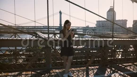 快乐的白种人女孩在电话中交谈站在令人惊叹的布鲁克林大桥附近在美丽的夏日4K的纽约景色中视频的预览图