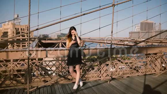 美丽的欧洲女性博主站在纽约布鲁克林大桥使用智能手机购物应用程序在一个阳光明媚的4K天视频的预览图