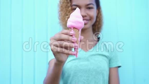 快乐的黑人女孩展示了粉红色融化的冰淇淋然后开始在蓝色的墙壁背景下吃它视频的预览图