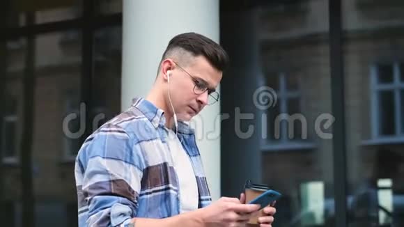 城市街道上的一次性纸杯上有智能手机喝咖啡的人的肖像英俊的年轻人视频的预览图