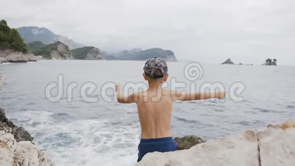 后景可爱的孩子举起双手站在海边的岩石上快乐的男孩高高举起双手视频的预览图