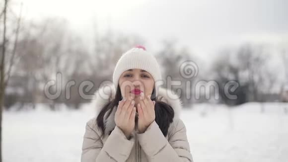 布吕内特的头发年轻女性的肖像粉红色的嘴唇穿着米色的羽绒大衣和帽子在白雪皑皑的中央公园里传递着一个空气的吻视频的预览图