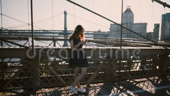 美丽的高加索女人站在布鲁克林大桥使用智能手机信使应用程序纽约惊人的天际全景4K视频的预览图