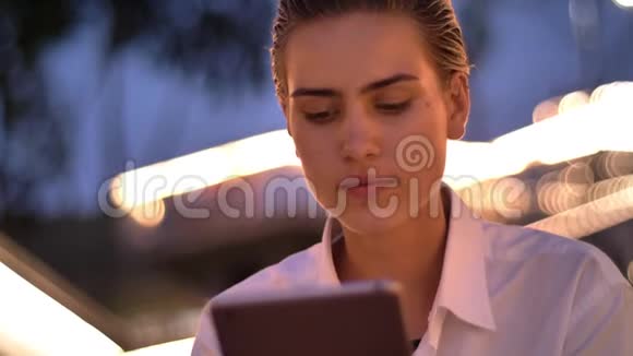 年轻的现代女性坐在梯子上晚上敲着平板电脑背景灯模糊视频的预览图